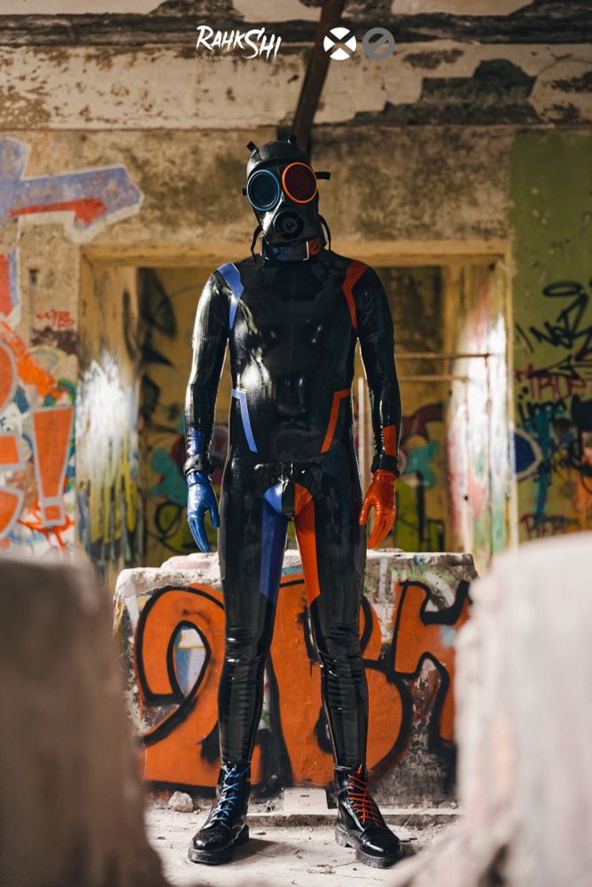 Rubber covered drone in black with blue and red stripes wearing a S10 mask with blue and red accents in the middle of a derelict building 