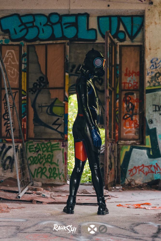 Wide shot of a derelict construction with an opening to the green outside.
In the middle stand a drone covered in black latex with blue and red stripes, back against us, turning to look at the camera mysteriously