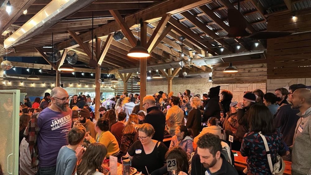 Large crowd, seated and standing, in a covered patio with lights.
