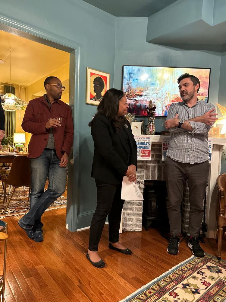 Rashida and Daniel are standing at the front of a room and looking at each other. Another man is in the background, in a doorway to a dining area.