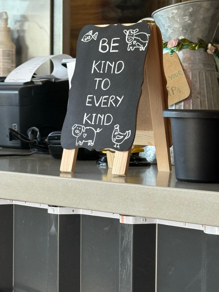 The front counter at a restaurant focused on a small sign on a mini easel that reads: Be kind to every kind