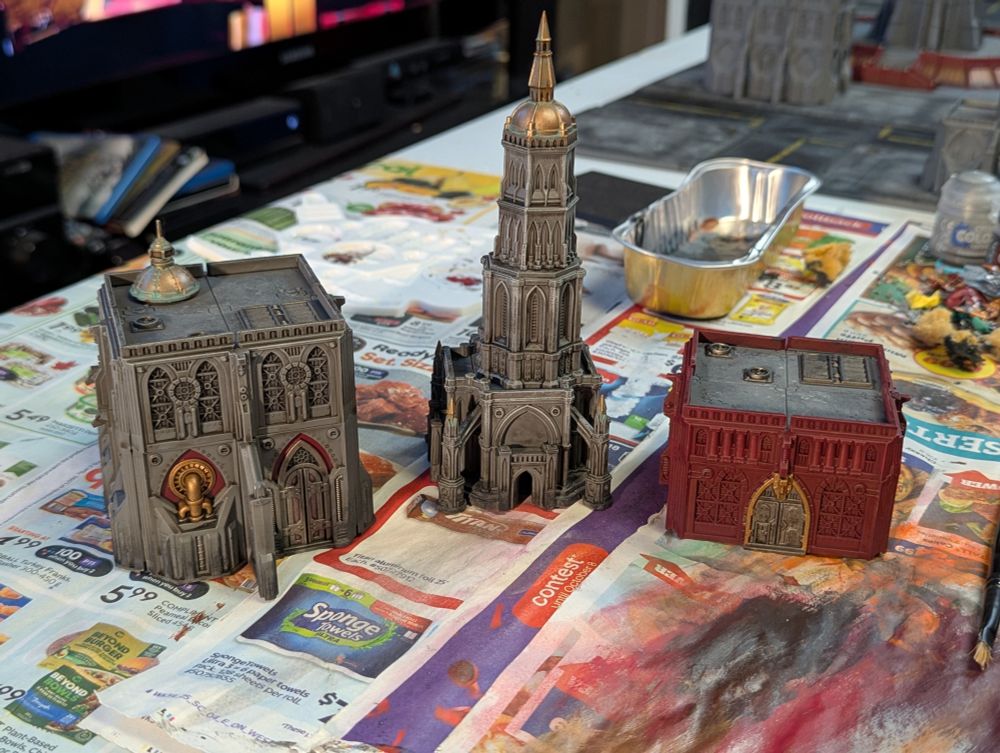 Three medium sized buildings scaled for the tabletop game Legions Imperialis sit on a painting table. On the left is a larger apartment building with a terrace and a grey with red accent paint job. The middle building is a shrine tower painted in a lighter grey with a copper spire edged with verdigris. The building on the right is a squat red apartment building with a grey roof.