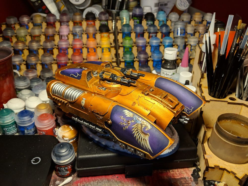 An Adeptus Custodes Coronus Grav-Carrier sits upon a painting desk, it has a rich gold and royal purple colour scheme. Viewed from the front right 3/4 perspective.