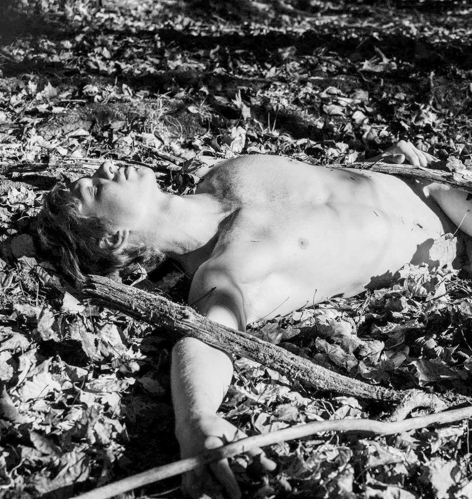 Black and white of a nude male figure lying unconscious on the forest floor amongst leaves and branches. Image is cropped from head to just below the waist.