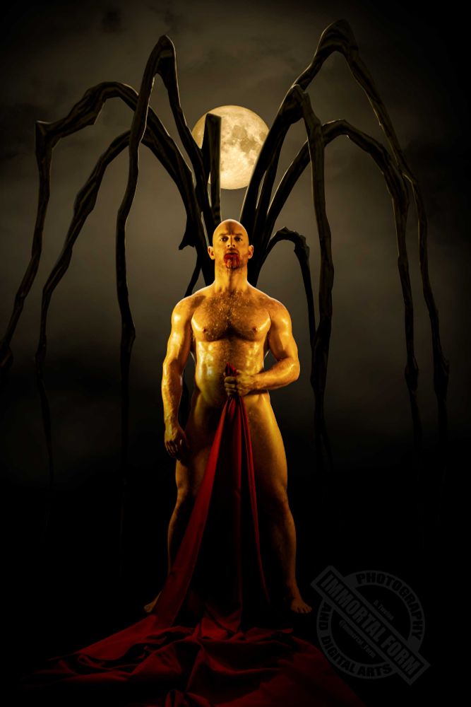 A young, nude muscled man with a hairy chest and bald head holding a red sheet in front of him by his left hand. Blood drips from his mouth. Behind him a giant black spider stands with the full moon in the sky. This is the full version of the photo showing the full nude figure of the man.