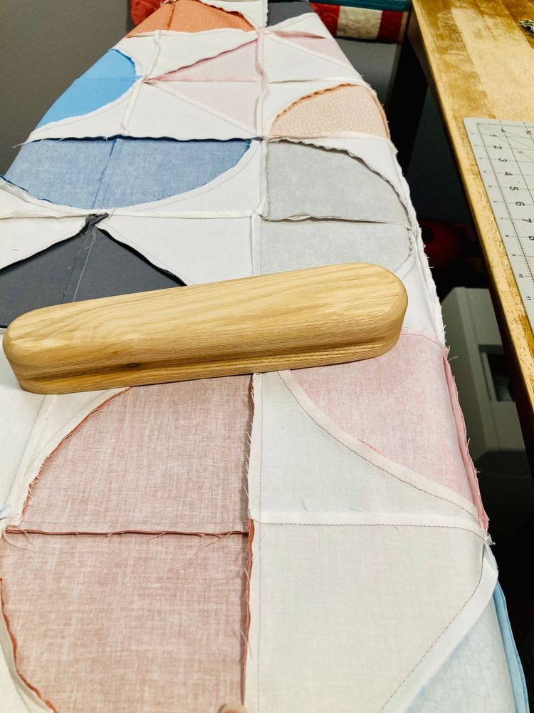 The back side of a colorful quilt top with several curved seams coming together. A small hardwood tailor's clapper is sitting on top of a newly pressed seam. 