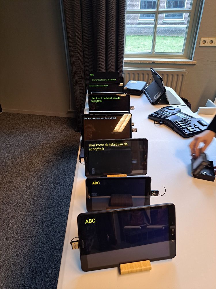 Op een witte tafel staan 7 tablets achter elkaar. Alle hebben zwarte achtergrond, de meeste met gele letters en 1 met witte letters.
Rechts achter is nog een velotype toetsenbord op tafel gelegd.