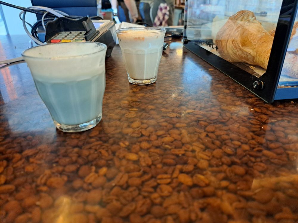 Links een klein limonadeglas met blauwe melkachtige vloeistof en opgeschuimde melk laag. Rechts iets meer naar achteren een lichtbruine versie van de vloeistof. Beide staan op een balie van koffiebonen onder glas.