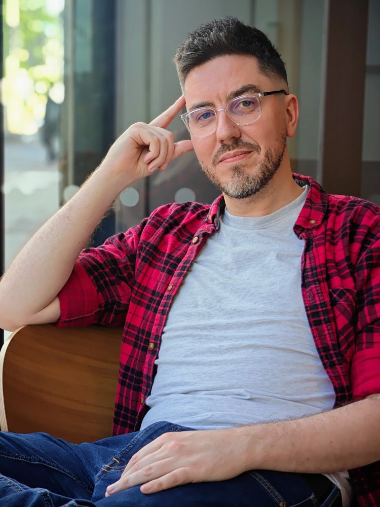 Hayden Scott smiling and looking at the camera. Red lumberjack shirt, relaxed pose, grey t-shirt. Photo from 2024.