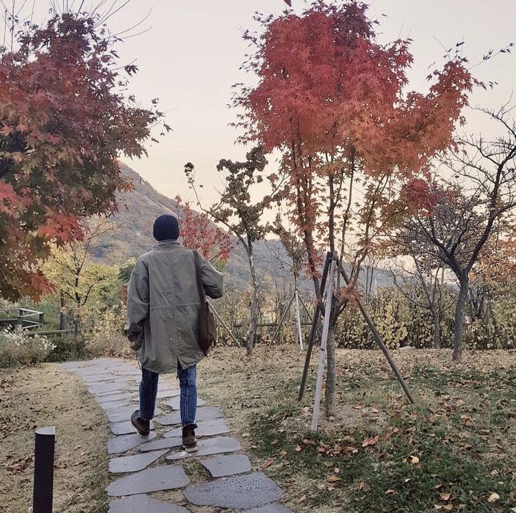 A picture of Namjoon from the back, striding down a stone path between autumnal trees. He’s wearing a beanie and a coat and jeans and boots. 