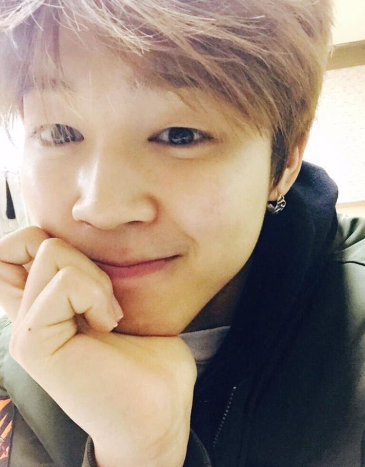 Blond Jimin looks very fondly at the camera. He is resting his chin in his right hand. 