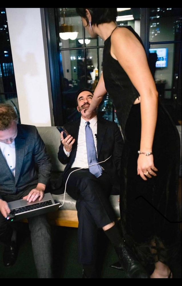 Zohran Mamdani is wearing a suit with a blue tie, holding a phone, and sitting down. His wife, Rama Duwaji, is standing over him, wearing a formal black two piece skirt set. Her right hand cradles Mamdani’s chin and he is looking up at her with great devotion. 
