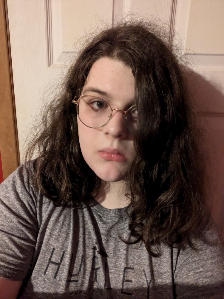 Myself in my bedroom. My face is a lot less serious but still looks dead inside. I have rose gold round framed glasses and shoulder-length hair. My face is generally softer, and my skin complexion is significantly lighter.