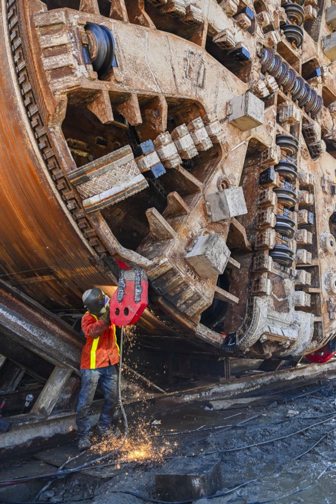 a large cutterhead on a tunnel boring machine stands tall preparing for its next project. in the lower left a worker is showering sparks onto the muddy ground as they grind a piece of the worn metal.