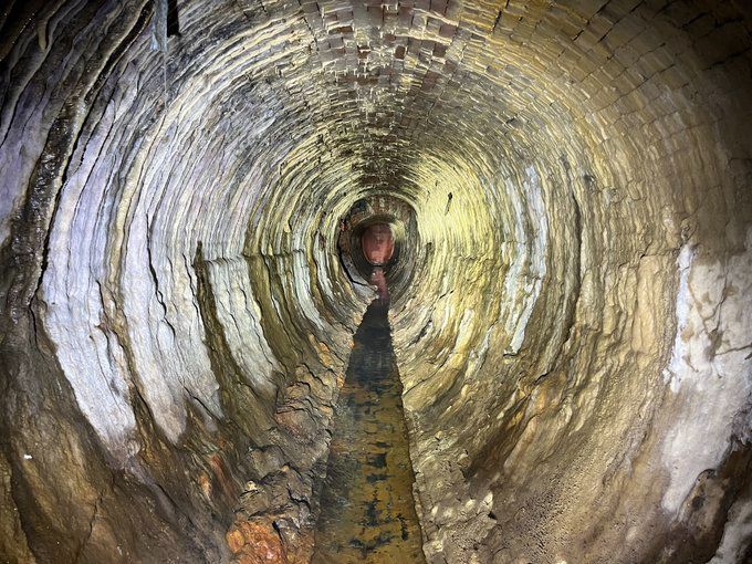 standing in sewer about 6 feet tall, old and lined with brick built in 1873. The brick is worn and covered in mineral deposits as the bottom has a small ankle-deep stream of water flowing away from the camera.
