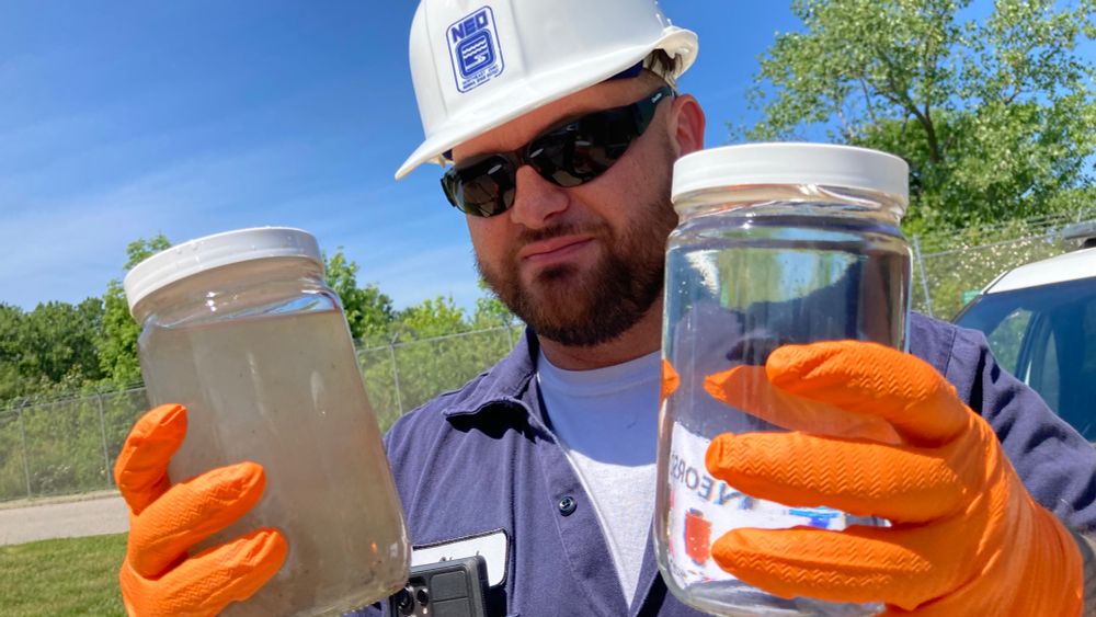 a man in a white hard hat holds two jars of water. he wears sunglasses. he looks very focused.