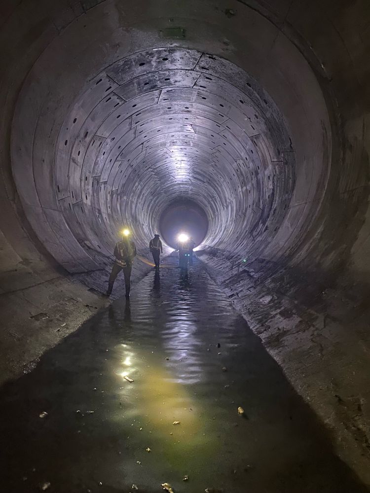 a long sewer tunnel, wide and tall and dark, with shin-deep brown water flowing at the bottom. in the distance are three figures, sewer workers, standing illuminated only by two hard hat head lamps.
