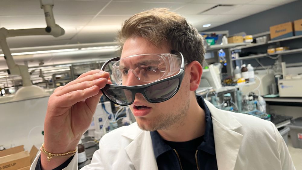 a man in a lab coat and safety glasses in a lab. he removes a pair of sunglasses to reveal safety glasses underneath.