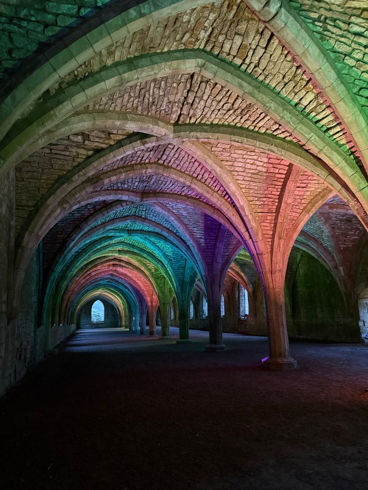 The celerium at Fountains Abbey, a well-preserved part of the ruin with a series of arches held up with pillars, with unglazed windows at the ends and along the sides. The arches are lit by different coloured lights, giving it a rainbow appearance.