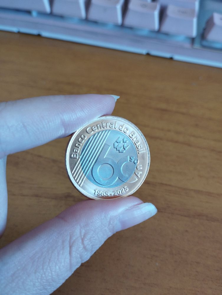 foto de dois dedos segurando uma moeda de um real, mostrando a parte de trás da moeda que apresenta um design comemorativo dos 60 anos do Banco Central do Brasil 