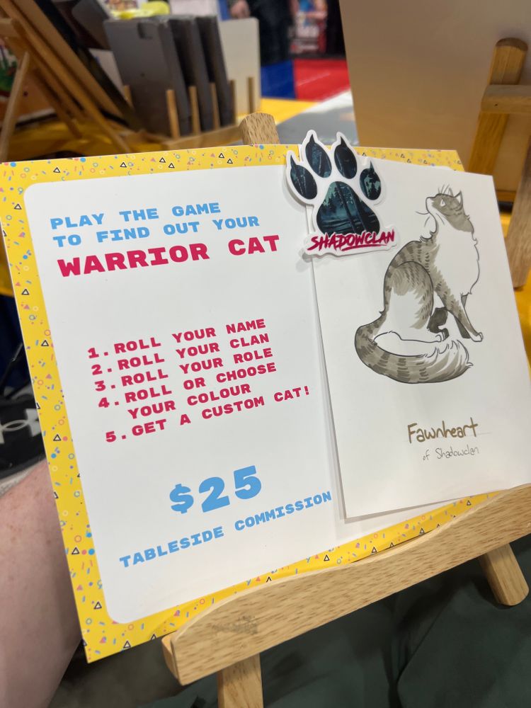 Commission information for the previous commission. It says “Play the game, find out your Warrior Cat” with a list of steps to generate a random cat from a table. A similar-looking cat called Fawnheart is attached to the sign - by pure chance, they rolled the same coloration. 
