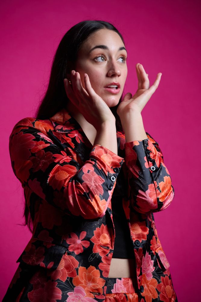 Photograph of Jordan wearing a pink, red, orange, and black suit over a crop-top black t-shirt. Behind her is a hot pink backdrop. She has her arms raised, and her hands positioned to either side of her jaw as she looks up and to photograph photographic right. Her eyebrows are raised and her lips parted.