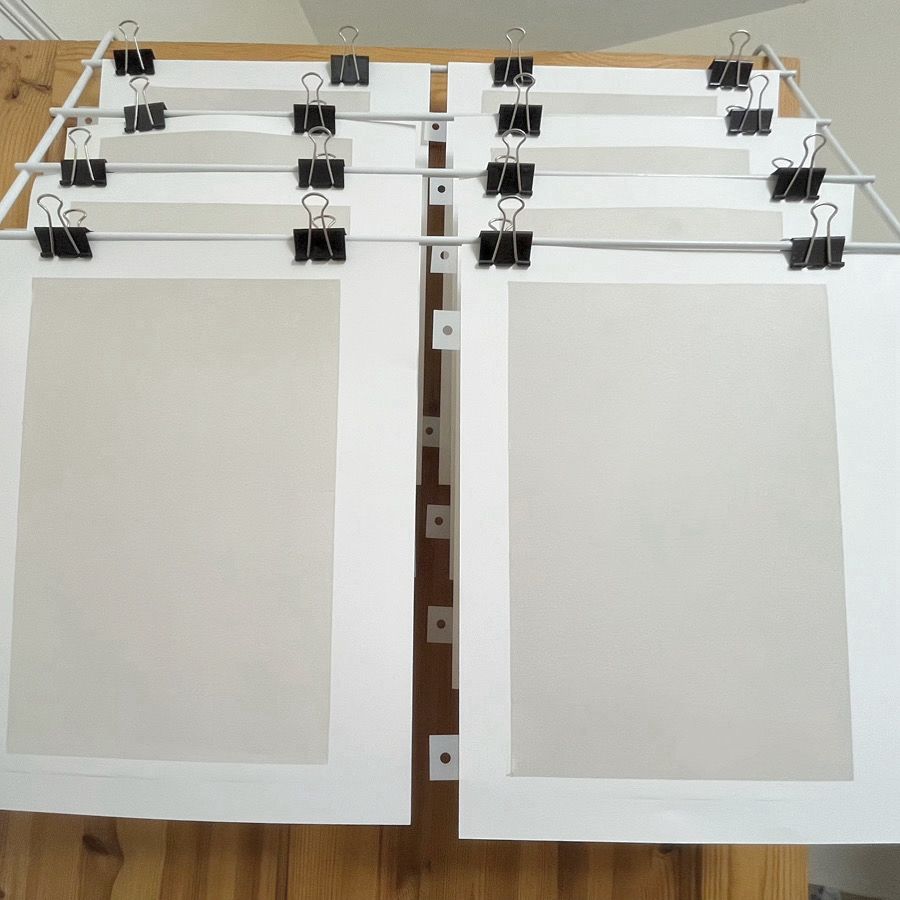 An over door clothes airer from which sheets of A4 printmaking paper, with a pale grey with a hint of brown solid ink base layer, are hanging by means of bulldog clips.