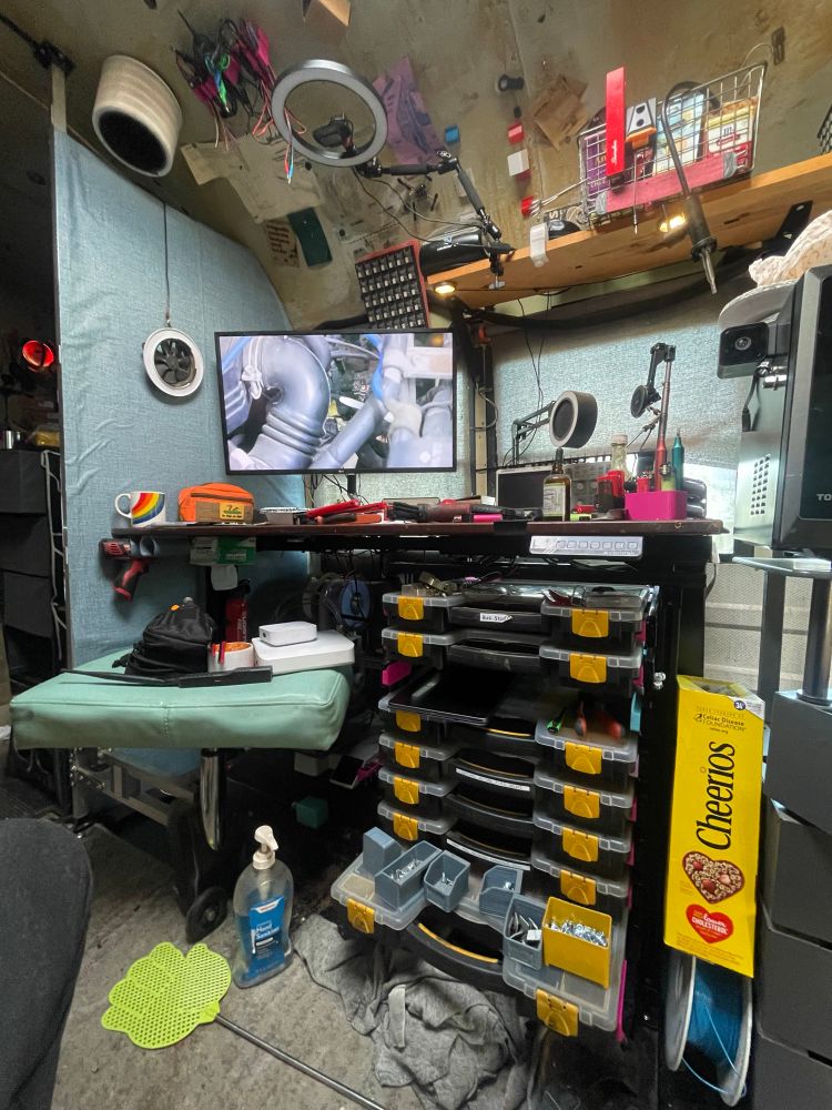 A lot happening around a desk in a school bus. There’s plentiful storage cases on sliders underneath a stool attached to the wall on top is a little messy with tools and a large monitor showing car engine parts. Lots of stuff is attached to the ceiling and walls of the bus
