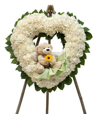 A heart shaped wreath of white flowers and green leaves on a stand, with a teddy bear attached to the wreath, and holding a sunflower. A green bow is seen under the teddy bears. (Pic from: flowersusa.net)