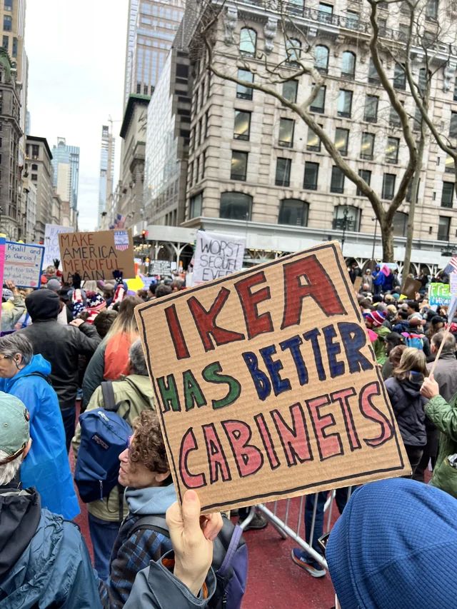 A cardboard sign seen at a protest alongside other signs; the sign in focus reads "Ikea has better cabinets".
