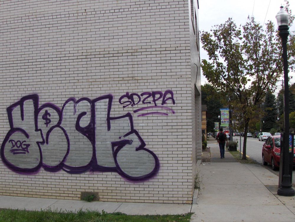 On the left is a large graffiti tag painted on the wall of a building.  On the right is a sidewalk lined with street lamps and small trees.  A person can be seen walking in the distance.