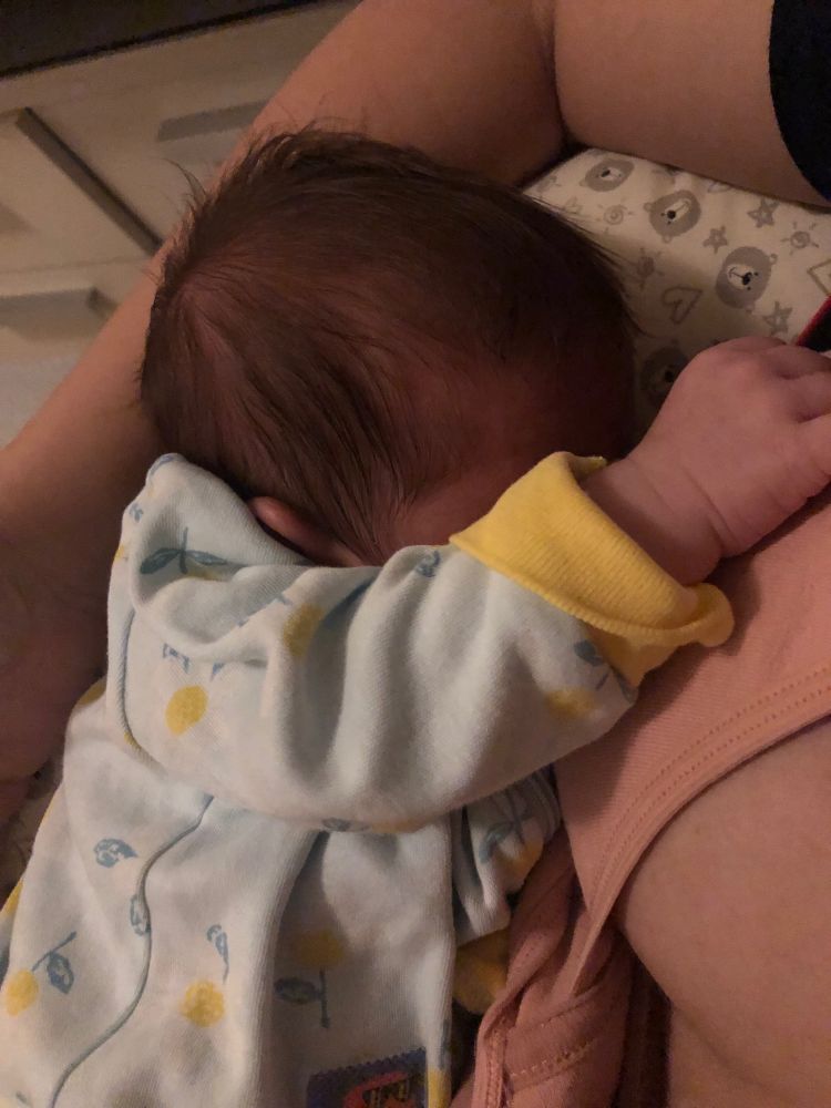 Bebê vestindo um macacão azul claro com elástico na manga amarelo, com desenhos de margaridas, escondendo o rosto da luz com o braço enquanto mama