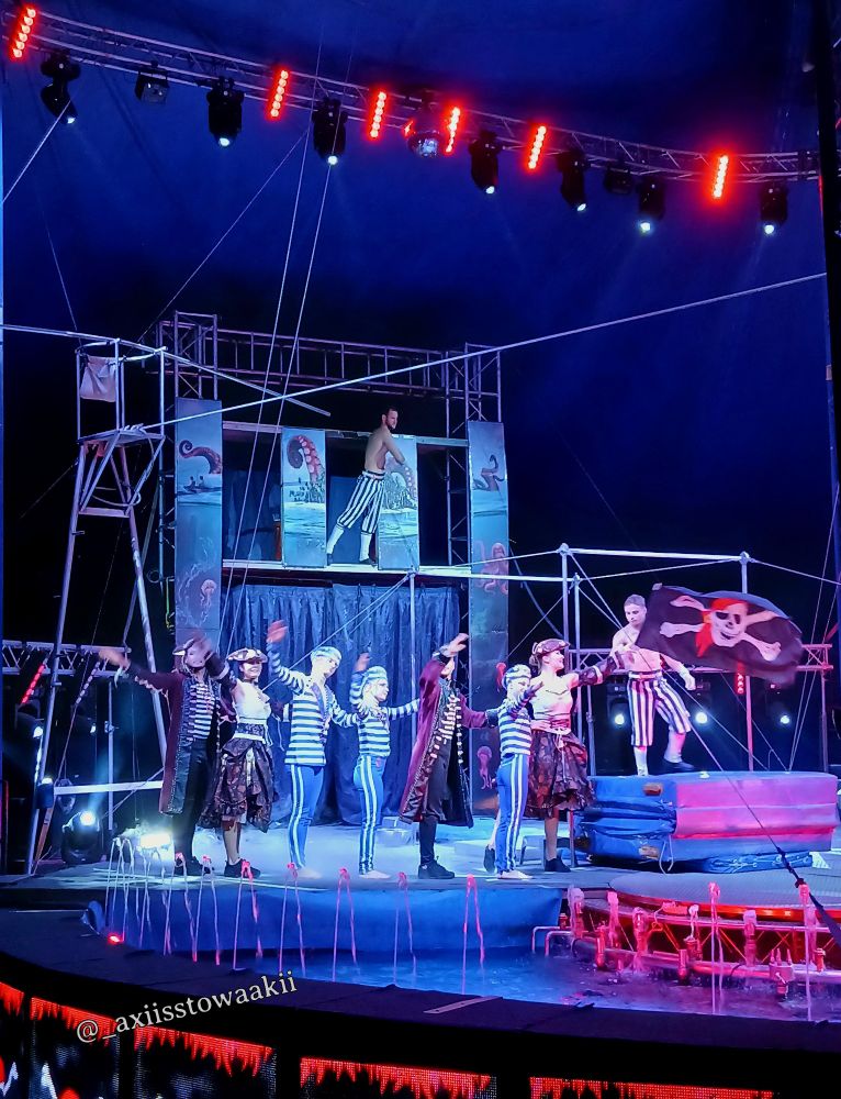 Zirkusbühne mit Artisten in gestreiften Kostümen und Piratenoutfits, beleuchtet von roten und weißen Scheinwerfern unter einem blauen Zirkuszelt.