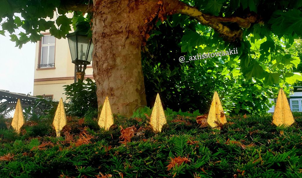Sechs goldene Zaunspitzen die durch eine Hecke stoßen, im Hintergrund ist noch ein Baum und eine Laterne zu sehn. 