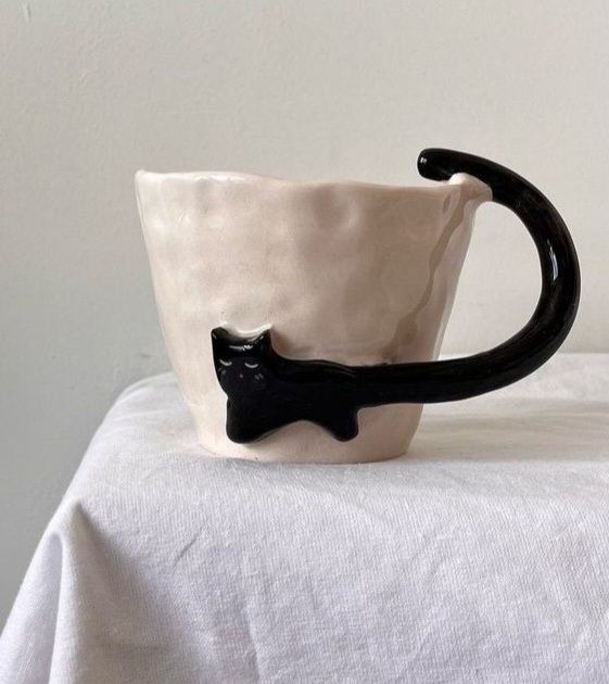 Eine weiße Tasse mit einer schwarzen Katze, deren Schwanz der Henkel ist. 
Netzfund