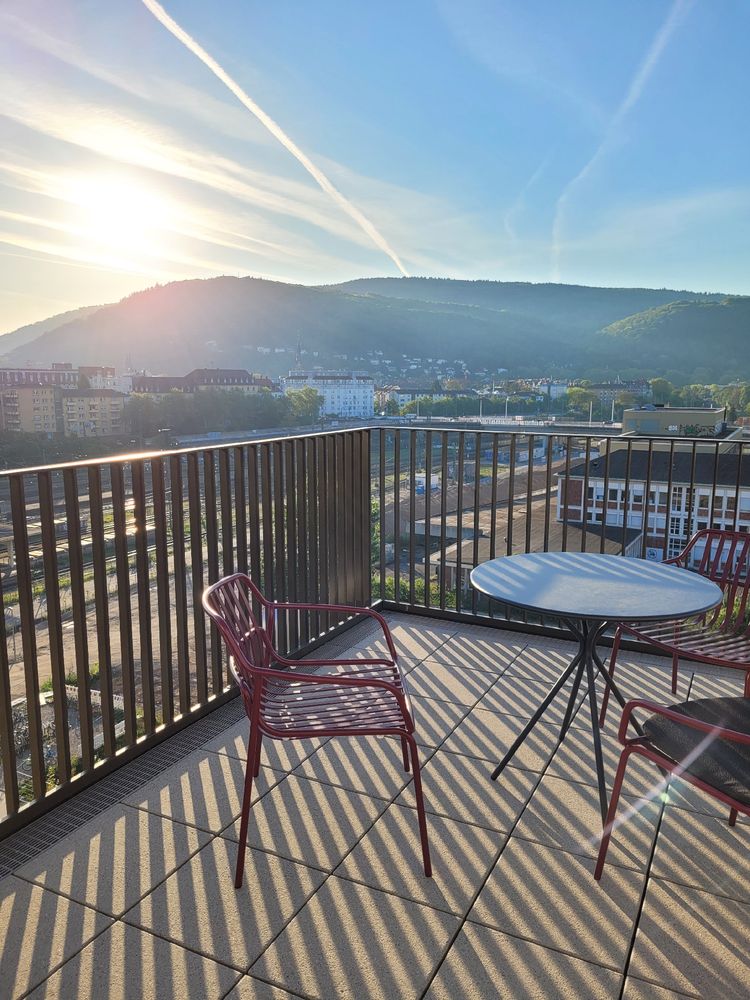 Teil einer Dachterrasse mit Stühlen und Tisch aus Metall.. Am Horizont geht die Sonne hinter einem Berg auf. 