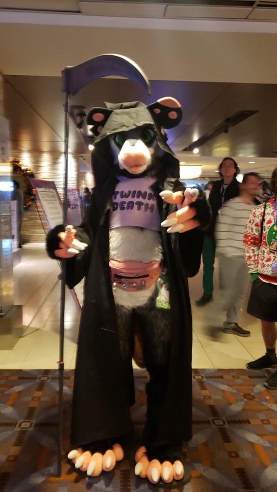Opossum fursuiter wearing a grim reaper hood and holding a scythe. They are also wearing a crop top shirt displaying the words "Twink Death" and fanny pack with enamel pins on it