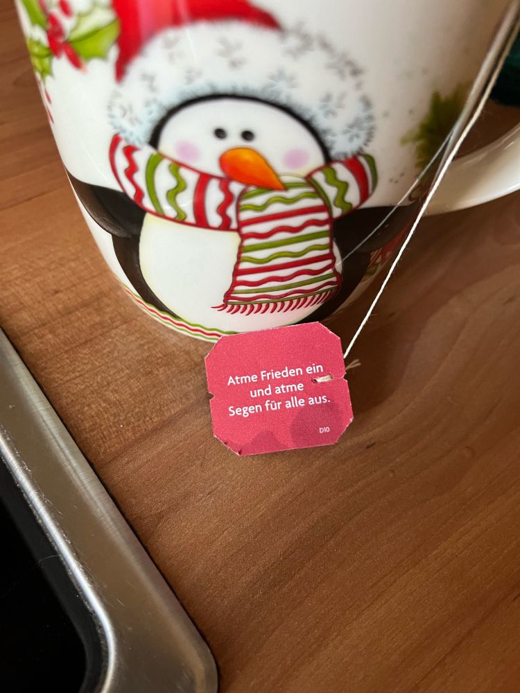 Ein Pinguinbecher steht auf Holzuntergrund. Auf dem Teebeutelschildchen, das über den Becherrand hängt, steht: „Atme Frieden ein und atme Segen für alle aus.“