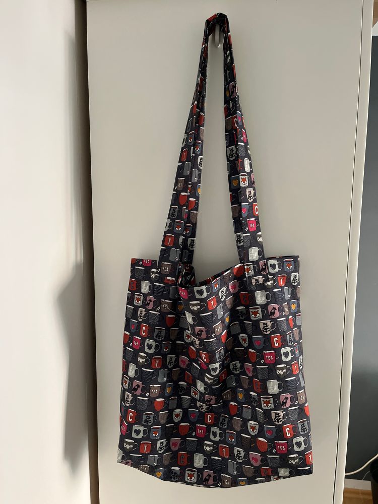 Ein selbstgenähter Tragebeutel mit vielen bunten Bechern auf grauem Grund hängt an der Seitenwand eines weißen Schranks.