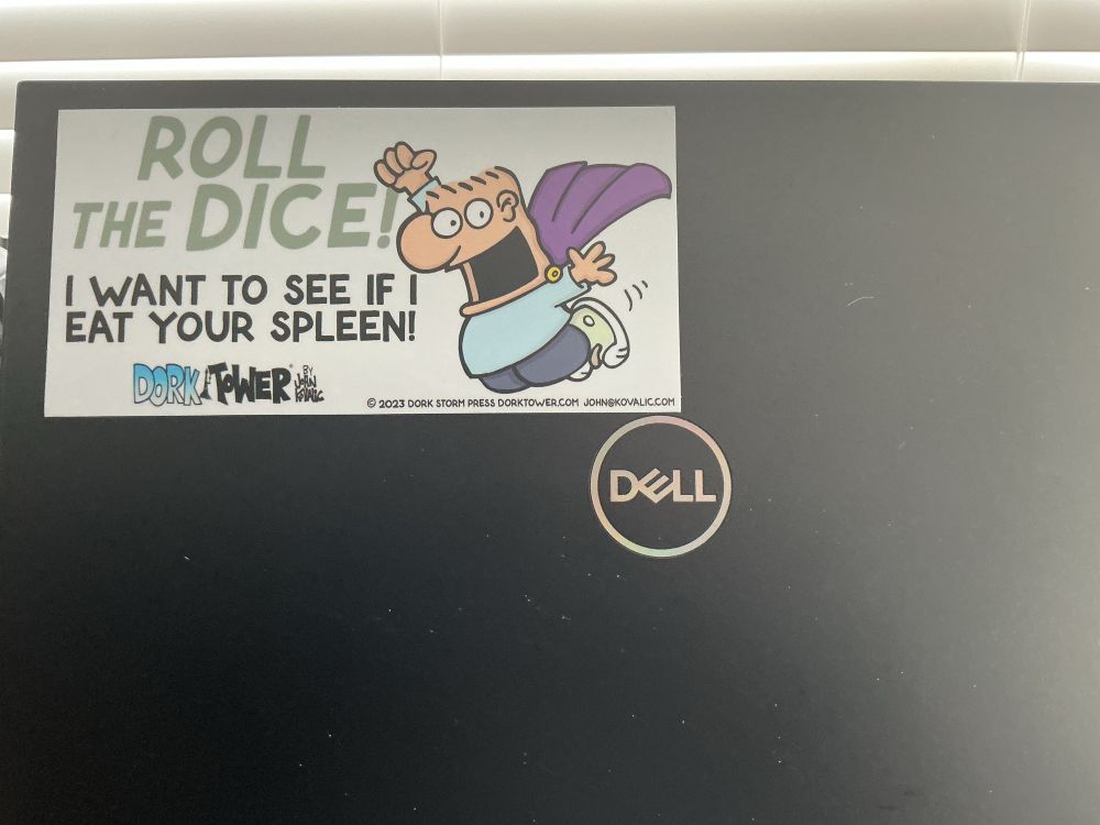 The back of my laptop with a bumper sticker showing Igor from Dork Tower John’s web cartoon. Igor is short man, flat head with a large expressive mouth. He is shown jumping while wearing a cape and saying “Roll the dice! I want to see if I eat your spleen.”