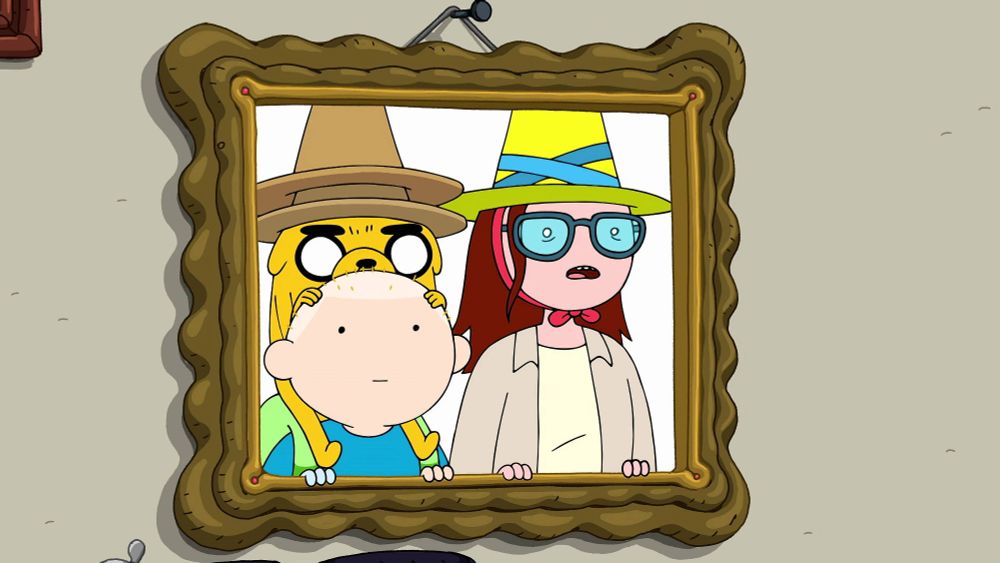 Jermaine wears 2 pointy hats while getting a piggy back ride from a bald Finn, both of them next to Betty as they look through a photo frame.