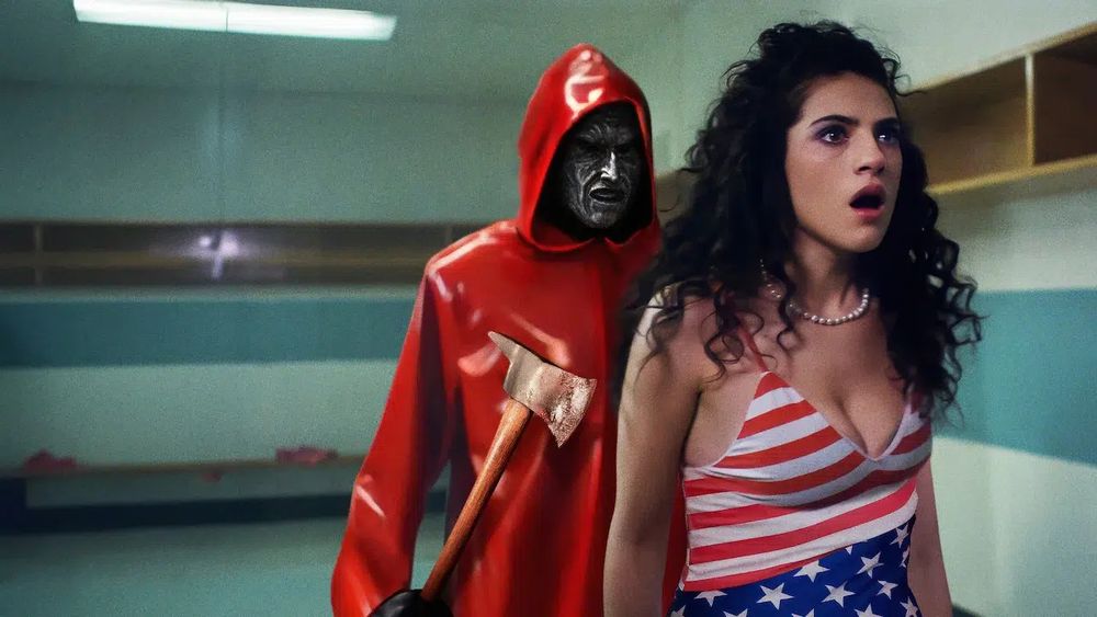 One of the prom queen candidates in an American flag swimsuit. The slasher of the film, in a red rubber coat, mask, and a hatchet, is looming behind her as she looks in the bathroom mirror.