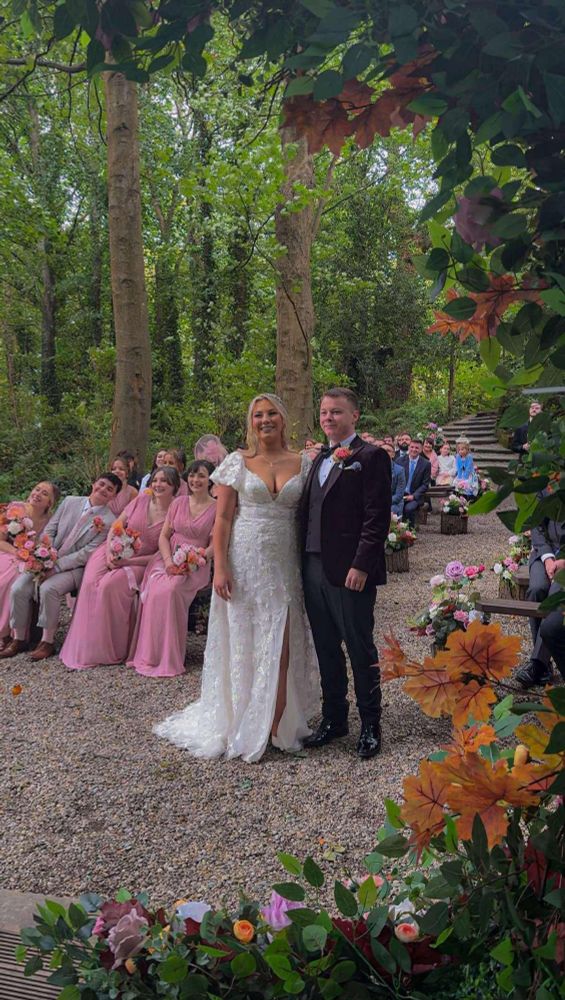 Bride and groom standing in a forest ceremony clearing. To the brides left three bridesmaids and the man of honour are seared and leaning into the shot.