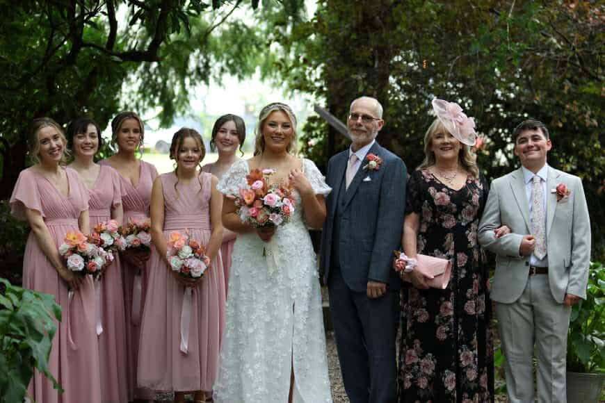 5 bridesmaids in pink stand to the left of the bride in white. Father and mother of the bride and man of honour to the brides right.