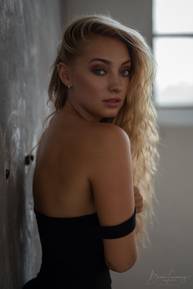 Eine junge Frau mit langen, welligen blonden Haaren steht seitlich an einer grauen Wand und blickt über die Schulter in Richtung Kamera. Sie trägt ein schulterfreies schwarzes Kleid, das leicht herabgerutscht ist. Weiches Tageslicht fällt durch ein Fenster im Hintergrund und beleuchtet ihr Gesicht und ihre Schultern, wodurch eine ruhige, intime Stimmung entsteht.