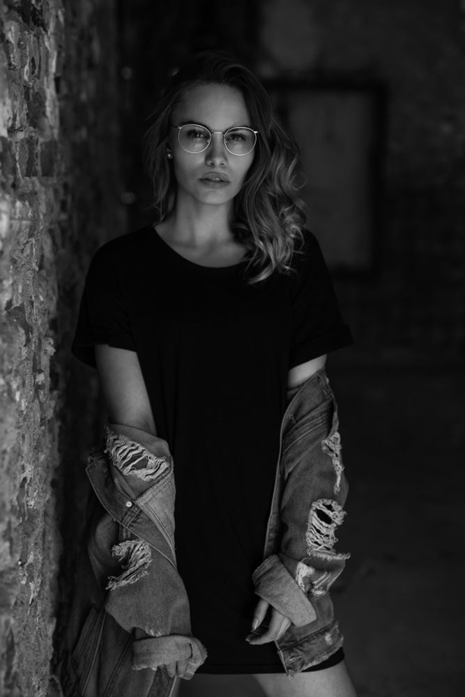 Schwarzweiß-Porträt einer jungen Frau mit runder Brille. Sie steht an einer rauen Steinwand in einem schmalen Gang, blickt ruhig in die Kamera. Sie trägt ein schlichtes dunkles Kleid und eine offen getragene, zerrissene Jeansjacke, die locker von den Schultern hängt. Das Licht ist weich, der Hintergrund unscharf.