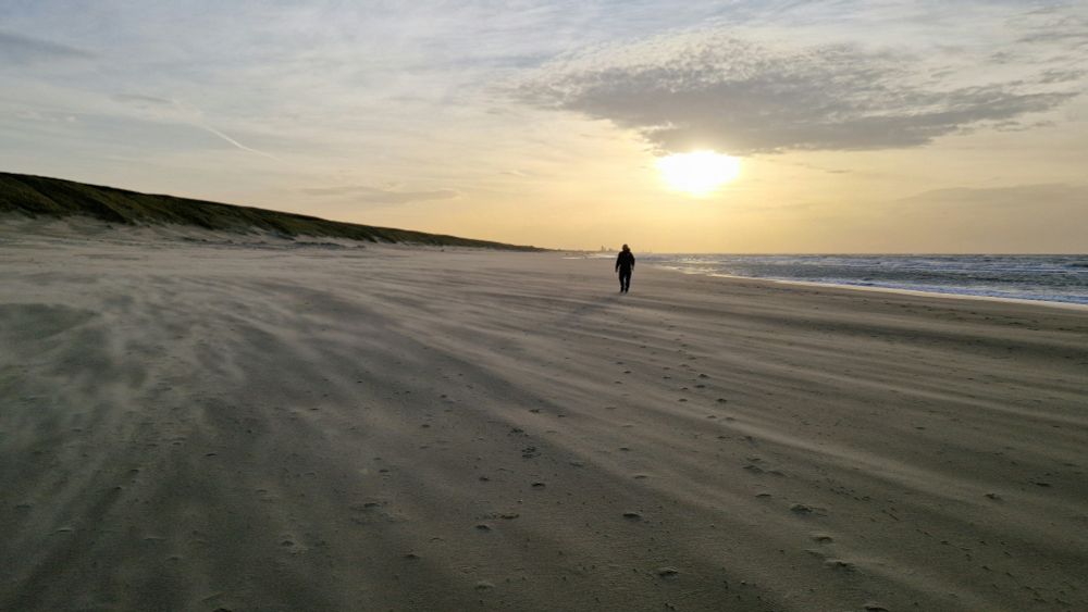 Op het strand, Wassenaarse Slag. Een eenzame wandelaar, de zon staat laag. De harde wind trekt stralen zand over de grond. 