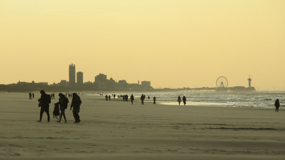 Zicht op Scheveningen vanaf Wassenaarse Slag. Tegenlicht, de lucht is oranje. Je ziet de pier en het reuzenrad. Veel wandelaars op het strand in silhouet. 