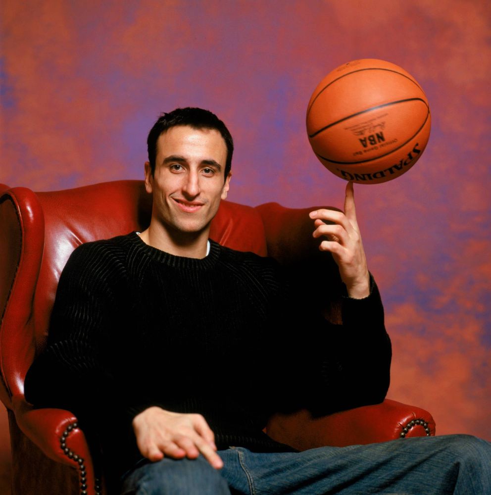 A young Manu Ginobili in a black Sweater and jeans sitting in a red leather chair in front of a fake sky background and twirling or balancing a basketball on his finger 