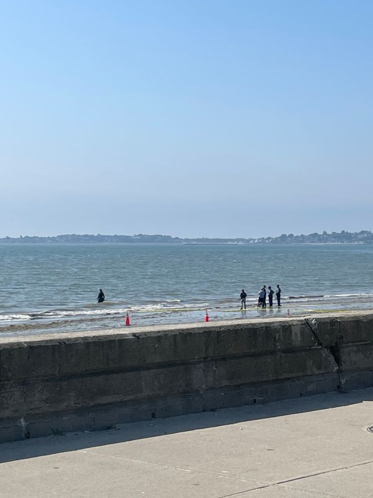 Photo of a sidewalk and sea wall at a public beach. In the background are Massachusetts State and local police on a cordoned off section of beach shore 